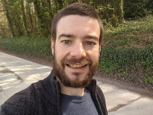 Man smiling outdoors, taking a selfie on a forest road with greenery in the background.