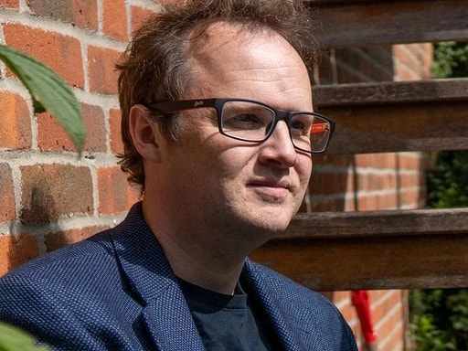 Man wearing glasses posing outside in the front of a brick wall.