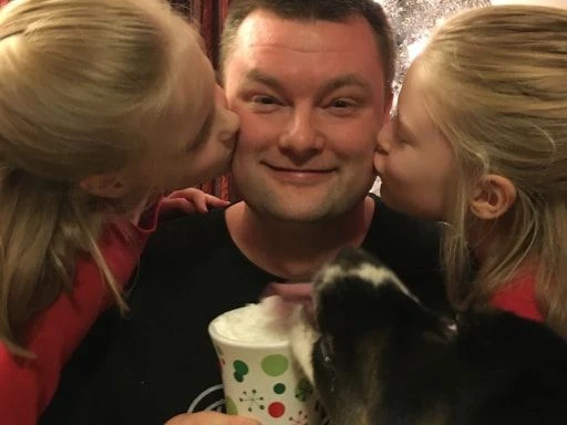 A joyful scene of a man holding a festive cup, being kissed on both cheeks by two girls while a dog licks the cup, suggesting a warm family moment.