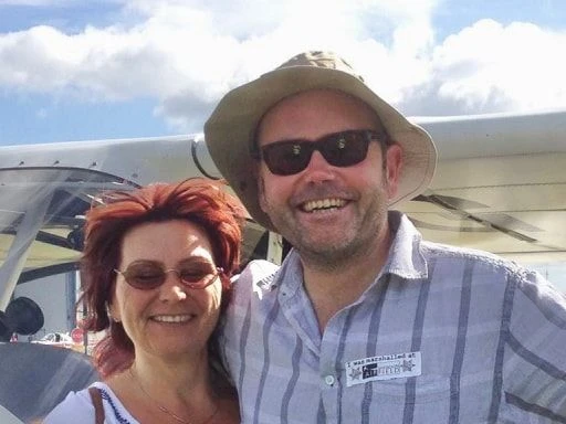 Man and woman smiling and wearing sunglasses, standing in front of a light aircraft.