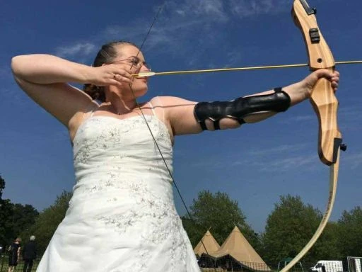 Woman in a wedding dress aiming a traditional wooden bow and arrow outdoors, with teepee tents in the background.