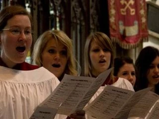 Group of choir singers dressed in traditional choir robes, singing from sheet music in a church setting.