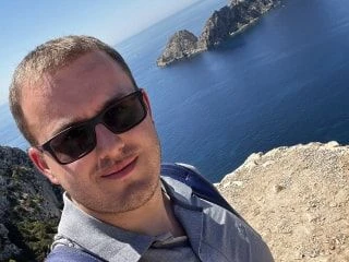 Man wearing sunglasses taking a selfie on a cliffside with a scenic ocean view and rocky islands in the background.