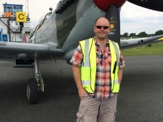 Man in a yellow safety vest smiling in front of a vintage aircraft on an airfield.